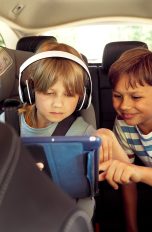 Reisen mit Kindern, Die besten Tipps für lange Autofahrten Zwei Kinder auf der Rückbank eines Autos schauen zusammen auf ein Tablet. Ein Kind trägt Kopfhörer.