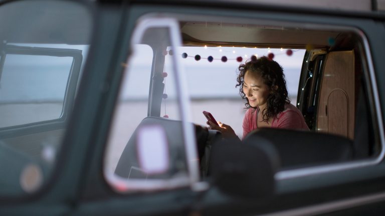 Blick durch das offene Fenster eines Campers, in dem eine Person an der offenen Seitent&uuml;r sitzt und auf ihr Handy schaut.