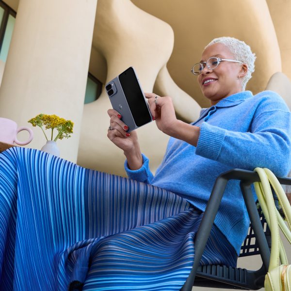 Eine Person sitzt vor einem futuristischen Gebäude auf einem Stuhl an einem Tisch und tippt auf ihrem aufgeklappten Google Pixel 10 Pro Fold.