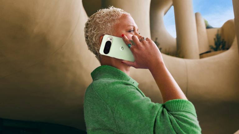 Eine Person steht vor einem futuristischen Gebäude und hält ein mintgrünes Google Pixel 10 Pro an ihr Ohr.