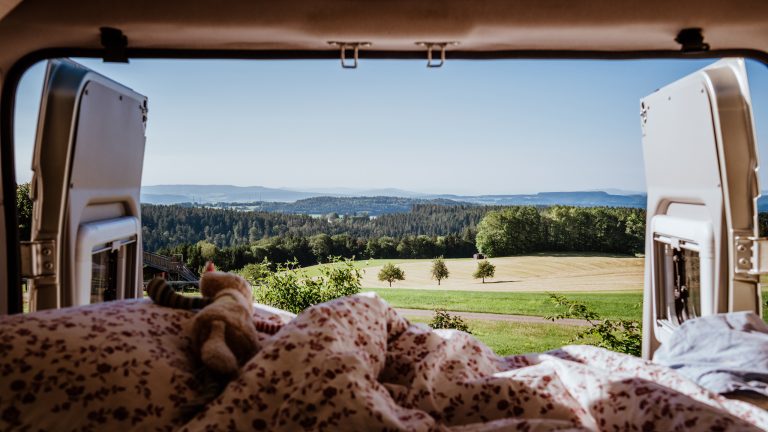 Blick aus dem Bett im ge&ouml;ffneten Heck eines Campervans auf Felder und dahinterliegenden Wald.