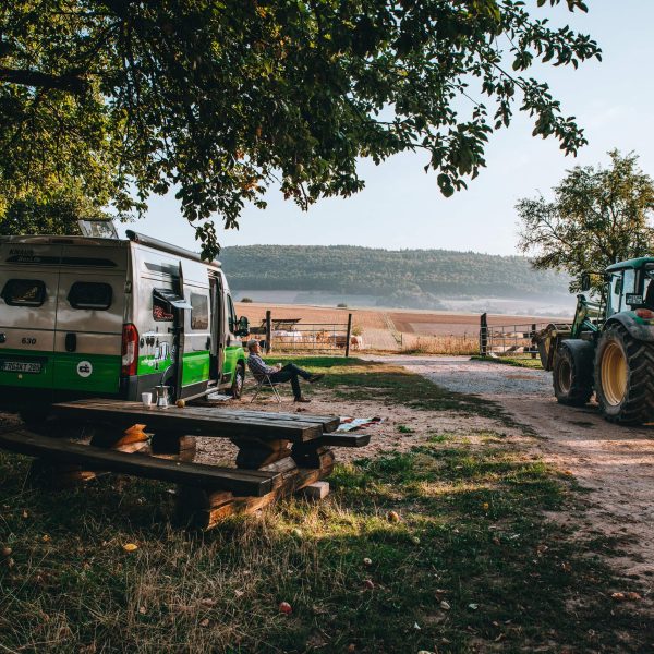 Ein Campervan steht neben einer Rastbank an einem Feldweg unter einem Baum. Auf dem Feldweg f&auml;hrt ein Traktor zu einem dahinterliegenden Feld.