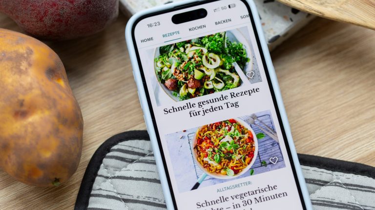 Ein Smartphone liegt neben Kartoffeln auf einer Holzoberfl&auml;che und einem Ofenhandschuh. Darauf ist die Startseite der Lecker-App mit schnellen Rezepten zu sehen.