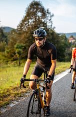 Zwei Personen fahren auf Rennr&auml;dern eine Landstra&szlig;e vor gr&uuml;nen B&auml;umen hinauf.