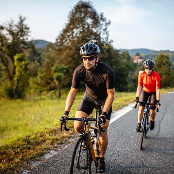 Zwei Personen fahren auf Rennr&auml;dern eine Landstra&szlig;e vor gr&uuml;nen B&auml;umen hinauf.