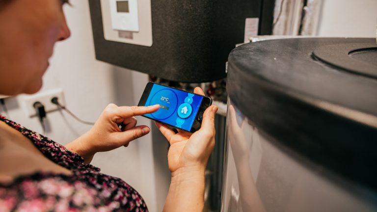 Eine Person steht im Heizungskeller. In der Hand h&auml;lt sie ein Smartphone, mit dem sie per App die Heizung steuert.