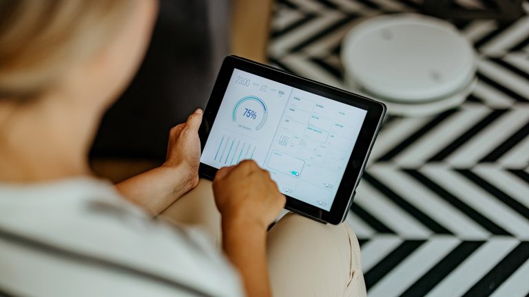 Aufnahme aus der Vogelperspektive einer Person, die ein Tablet in den H&auml;nden h&auml;lt auf dessen Display eine App zur Smarthome-Steuerung ge&ouml;ffnet ist.