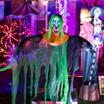 Ein schauriger Clown steht vor einem gruselig geschmückten Haus. Im Hintergrund ist eine weitere schaurige Figur zu sehen.