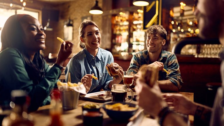 Mehrere Personen sitzen fr&ouml;hlich an einem Tisch in einem Pub und essen Burger und Pommes.
