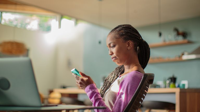 Eine Person sitzt an einem Schreibtisch in ihrem Zuhause und schaut auf ihr Smartphone.