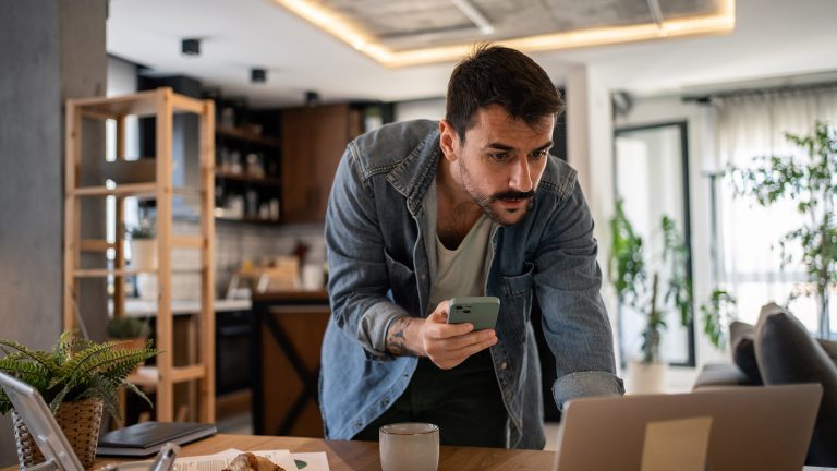 Eine Person beugt sich &uuml;ber ihren Wohnzimmertisch, auf dem ein Laptop steht. In den H&auml;nden h&auml;lt die Person ein Smartphone.