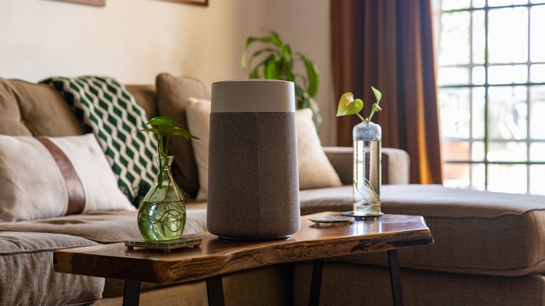 Ein Luftreiniger steht auf einem Couchtisch aus Holz. Daneben steht eine gr&uuml;ne Vase, im Hintergrund ist ein Sofa mit Zierkissen in Erdt&ouml;nen zu sehen.