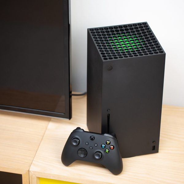 Foto einer Xbox Series X, die mit angelehntem Controller neben einem Fernseher steht.