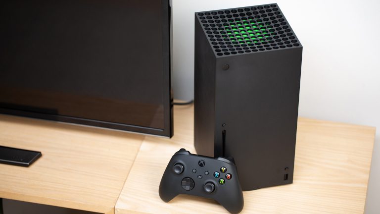 Foto einer Xbox Series X, die mit angelehntem Controller neben einem Fernseher steht.