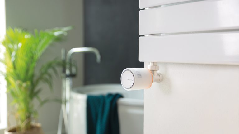 Nahaufnahme eines Netatmo-Thermostates, das an einer Badezimmer-Heizung angebracht ist. Im Hintergrund ist eine Badewanne zu erkennen.