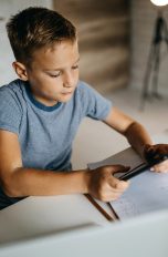Ein Kind sitzt mit einem Tablet in der Hand an einem Schreibtisch, auf dem ein Block liegt.