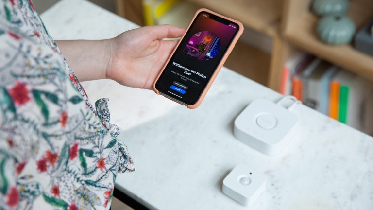 Eine Person hält ein Smartphone in der Hand, auf dem der Begrüßungstext der Philips-Hue-App zu sehen ist. Auf einem Tisch vor der Person liegen die Bridge und ein Bewegungsmelder.