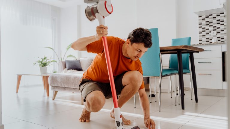 Eine Person sitzt in der Hocke. Mit der linken Hand wischt sie mit einem Tuch den Boden, in der rechten Hand hält sie ein kabelloses Kombi-Gerät.