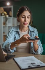 Eine Person sitzt an einem Schreibtisch. Vor ihr steht ein geöffneter Laptop, ein Klemmbrett mit Dokumenten liegt daneben. Über diese hält die Person ihr Smartphone.