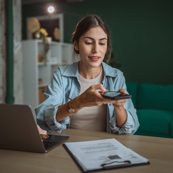 Eine Person sitzt an einem Schreibtisch. Vor ihr steht ein geöffneter Laptop, ein Klemmbrett mit Dokumenten liegt daneben. Über diese hält die Person ihr Smartphone.