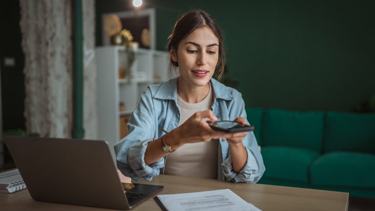 Eine Person sitzt an einem Schreibtisch. Vor ihr steht ein ge&ouml;ffneter Laptop, ein Klemmbrett mit Dokumenten liegt daneben. &Uuml;ber diese h&auml;lt die Person ihr Smartphone.