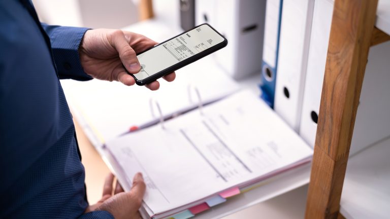 Nahaufnahme der Hand einer Person, die ein Smartphone &uuml;ber einen ge&ouml;ffneten Ordner mit Dokumenten, h&auml;lt.