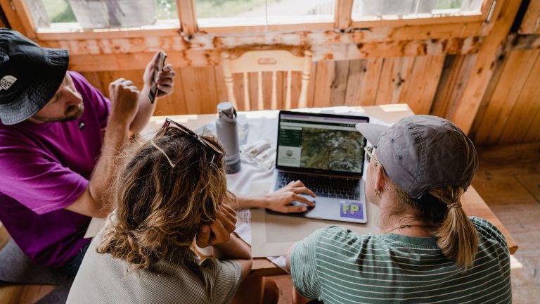 Drei Personen in Wanderkleidung sitzen an einem Tisch in einer H&uuml;tte vor einem Laptop und planen eine Bergtour.