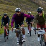Eine Gruppe von vier Gravelbiker*innen fährt auf einem Kiesweg. Im Hintergrund sind Berge zu sehen.