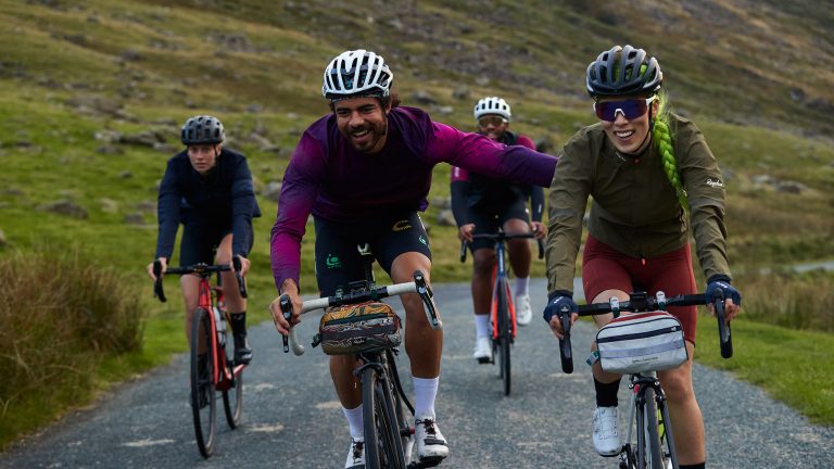 Eine Gruppe von vier Gravelbiker*innen f&auml;hrt auf einem Kiesweg. Im Hintergrund sind Berge zu sehen.