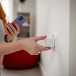 Nahaufnahme der Hand einer Person, die einen Smart Plug in eine Steckdose steckt. In der anderen Hand hält sie ein Smartphone.