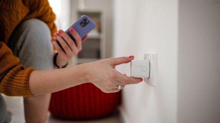 Nahaufnahme der Hand einer Person, die einen Smart Plug in eine Steckdose steckt. In der anderen Hand h&auml;lt sie ein Smartphone.