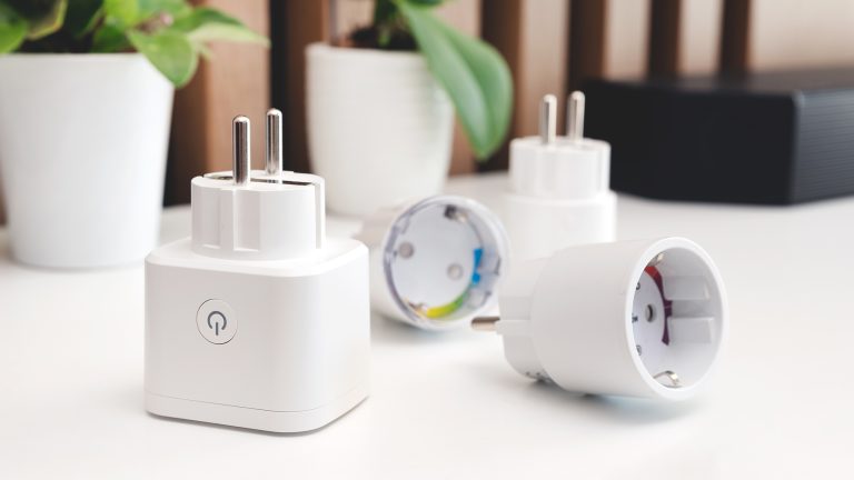 Zwei Smart Plugs liegen und zwei Smart Plugs stehen auf einem Tisch. Im Hintergrund stehen zwei Pflanzen.