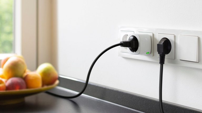 Zwei Smart Plugs stecken in einer Steckdose in der K&uuml;che. Im Hintergrund ist eine Obstschale zu sehen.