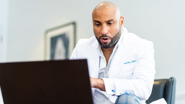 Eine Person in einem Arztkittel sitzt vor einem Laptop und diktiert etwas.