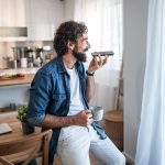 Eine Person lehnt an einem Esszimmertisch, während sie in der linken Hand ein Smartphone hält, in das sie spricht. In der rechten Hand hält die Person eine Tasse; im Hintergrund ist die Küche zu sehen.