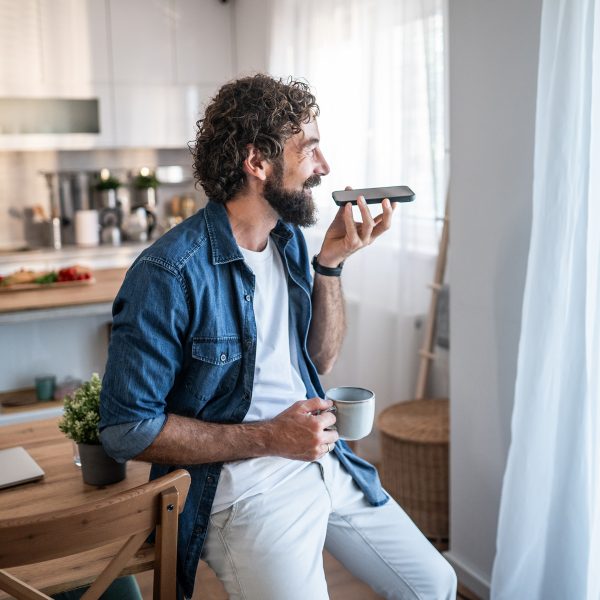Eine Person lehnt an einem Esszimmertisch, während sie in der linken Hand ein Smartphone hält, in das sie spricht. In der rechten Hand hält die Person eine Tasse; im Hintergrund ist die Küche zu sehen.