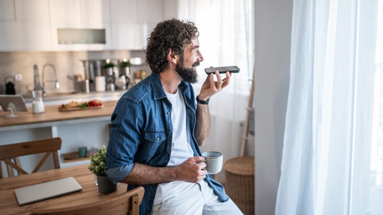 Eine Person lehnt an einem Esszimmertisch, während sie in der linken Hand ein Smartphone hält, in das sie spricht. In der rechten Hand hält die Person eine Tasse; im Hintergrund ist die Küche zu sehen.