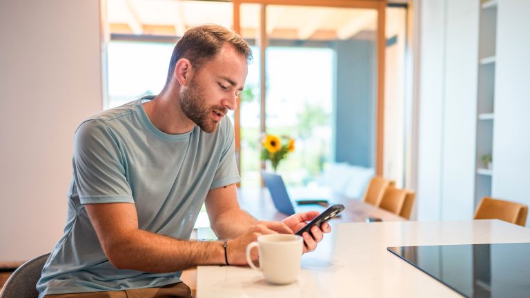 Eine Person sitzt an einem Küchentisch. Neben ihr steht eine Kaffeetasse; in den Händen hält sie ein Smartphone.