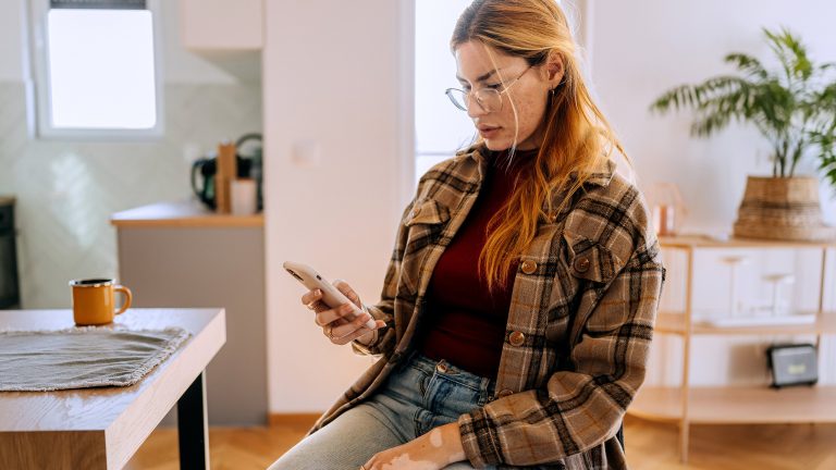 Eine Person sitzt auf einem Stuhl am Esszimmertisch. In der rechten Hand hält sie ein Smartphone.