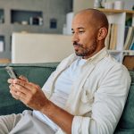 Eine Person sitzt auf einem Sofa im Wohnzimmer und schaut auf ihr Smartphone, das sie in beiden Händen hält.