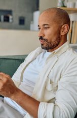Eine Person sitzt auf einem Sofa im Wohnzimmer und schaut auf ihr Smartphone, das sie in beiden H&auml;nden h&auml;lt.