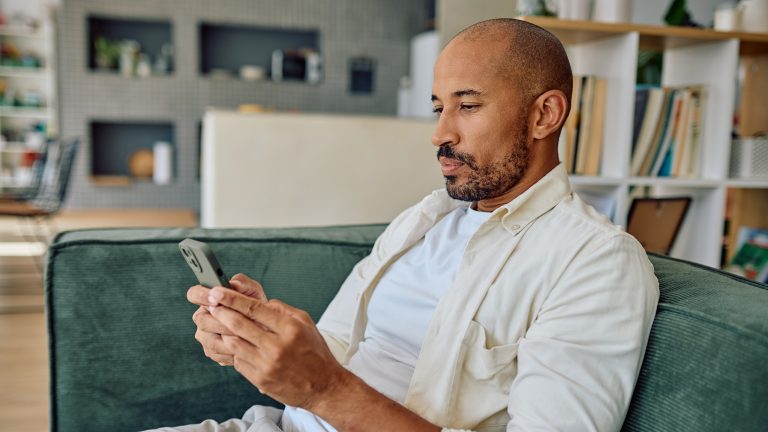 Eine Person sitzt auf einem Sofa im Wohnzimmer und schaut auf ihr Smartphone, das sie in beiden Händen hält.