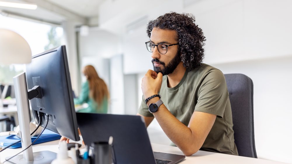 Eine Person sitzt in einem Großraumbüro vor einem Laptop und einem Bildschirm, in den sie fragend hineinschaut.