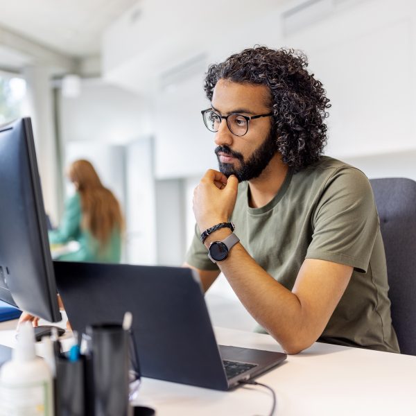 Eine Person sitzt in einem Großraumbüro vor einem Laptop und einem Bildschirm, in den sie fragend hineinschaut.