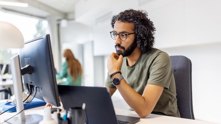 Eine Person sitzt in einem Großraumbüro vor einem Laptop und einem Bildschirm, in den sie fragend hineinschaut.