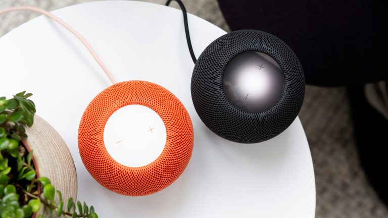 Zwei HomePod minis in orange und schwarz stehen auf einem weißen Beistelltisch neben einer Pflanze.
