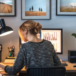 Eine Person von hinten, die an einem Schreibtisch vor einem Bildschirm und einem Tablet sitzt und etwas in einem Notizbuch nachliest.