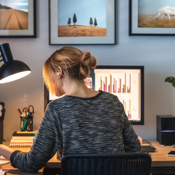 Eine Person von hinten, die an einem Schreibtisch vor einem Bildschirm und einem Tablet sitzt und etwas in einem Notizbuch nachliest.