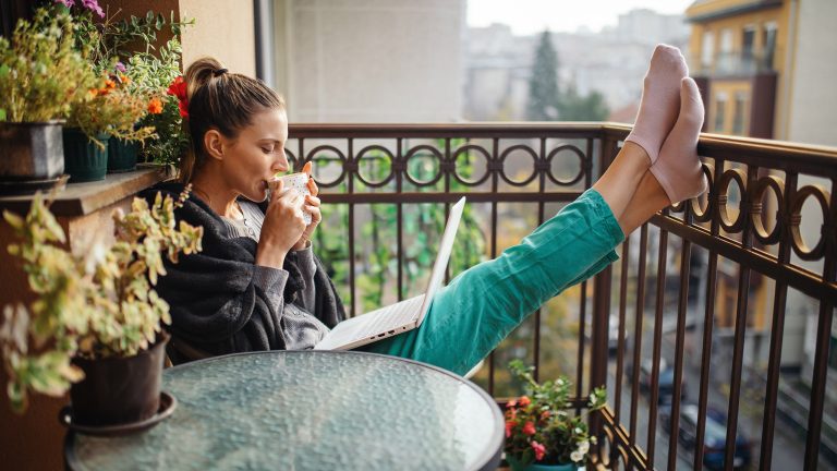 Eine Person sitzt mit einem Laptop auf dem Schoß auf einem Balkon und stützt ihre Füße ans Geländer. Sie trinkt gerade aus einer Tasse in beiden Händen.