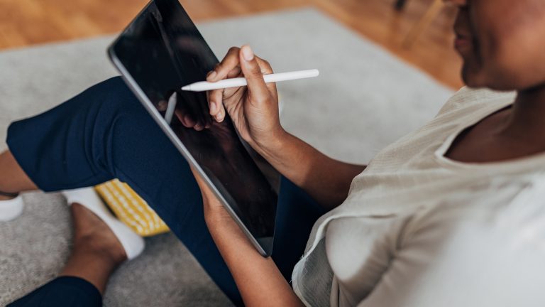 Vogelperspektive auf eine Person, die im Schneidersitz auf dem Boden sitzt und ein Tablet auf ihrem Schoß mit einem Stylus-Stift bedient.
